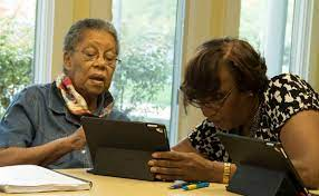Two older female adults seated at a table with one explaining something on a tablet.
